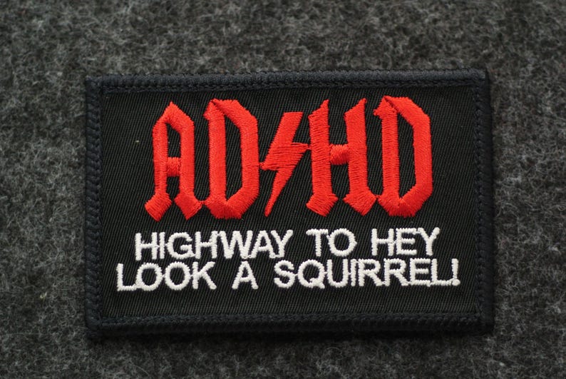 May include: Black rectangular patch with red embroidered text "ADHD" and a lightning bolt, above "HIGHWAY TO HEY LOOK A SQUIRREL!" in white. The patch has a black border and is set against a gray background.