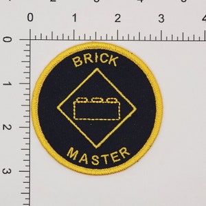 May include: A black and yellow embroidered patch with a diamond shape in the center. The diamond has a small brick design inside. The text "BRICK MASTER" is written around the outside of the patch.