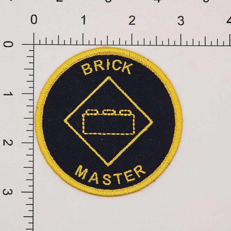 May include: A black and yellow embroidered patch with a diamond shape in the center. The diamond has a small brick design inside. The text "BRICK MASTER" is written around the outside of the patch.