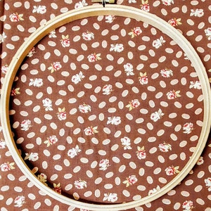 Puede incluir: Un aro de bordado de madera con un fondo de tela marr&oacute;n con un patr&oacute;n floral blanco. El aro es redondo y tiene un di&aacute;metro de aproximadamente 25 cm.