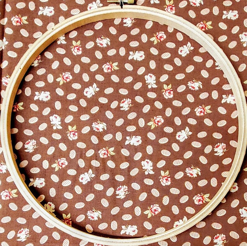 Puede incluir: Un aro de bordado de madera con un fondo de tela marr&oacute;n con un patr&oacute;n floral blanco. El aro es redondo y tiene un di&aacute;metro de aproximadamente 25 cm.