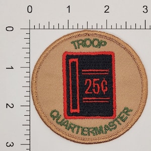 Pu&ograve; includere: Una toppa circolare color beige con testo ricamato in verde che recita "TROOP QUARTERMASTER" attorno al bordo. Al centro c'&egrave; un'immagine ricamata in nero e rosso di un libro mastro con il testo "25&cent;" in rosso.