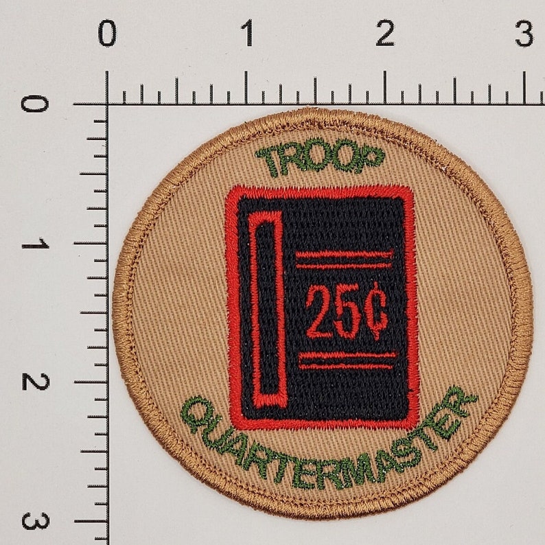 Pu&ograve; includere: Una toppa circolare color beige con testo ricamato in verde che recita "TROOP QUARTERMASTER" attorno al bordo. Al centro c'&egrave; un'immagine ricamata in nero e rosso di un libro mastro con il testo "25&cent;" in rosso.