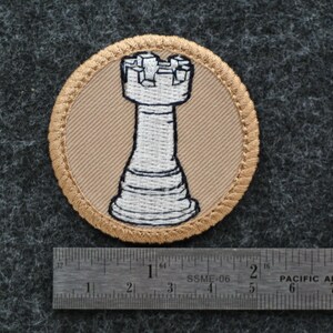 Op de afbeelding: Ronde geborduurde patch met een witte toren schaakstuk op een beige achtergrond, omlijst door een gouden rand. De patch heeft een diameter van ongeveer 6,35 cm. De achtergrond is een donkergrijze, getextureerde stof.