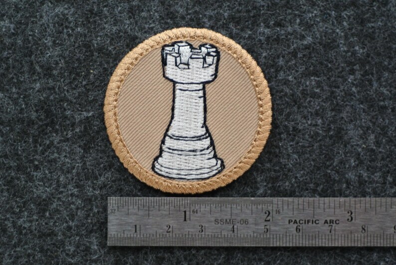 Op de afbeelding: Ronde geborduurde patch met een witte toren schaakstuk op een beige achtergrond, omlijst door een gouden rand. De patch heeft een diameter van ongeveer 6,35 cm. De achtergrond is een donkergrijze, getextureerde stof.