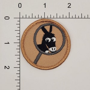 Op de afbeelding: Een bruine stoffen patch met een grijze cirkel en een zwarte ezel erin. De ezel heeft witte ogen en een witte neus.