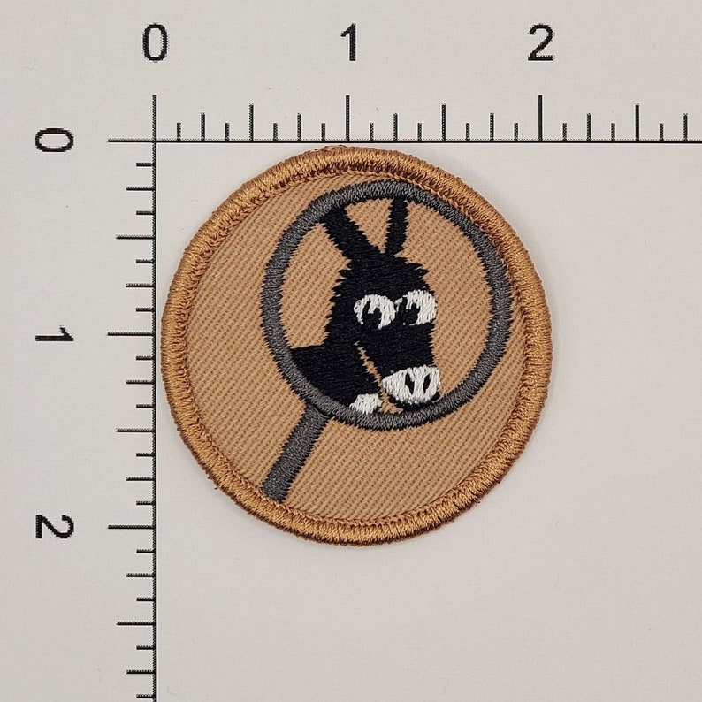 Op de afbeelding: Een bruine stoffen patch met een grijze cirkel en een zwarte ezel erin. De ezel heeft witte ogen en een witte neus.