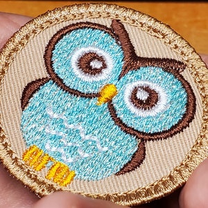 May include: Embroidered patch featuring a cartoon owl. The owl has a teal body, brown accents, and white eyes. The patch is round with a tan background and a gold border. The owl's beak is yellow.