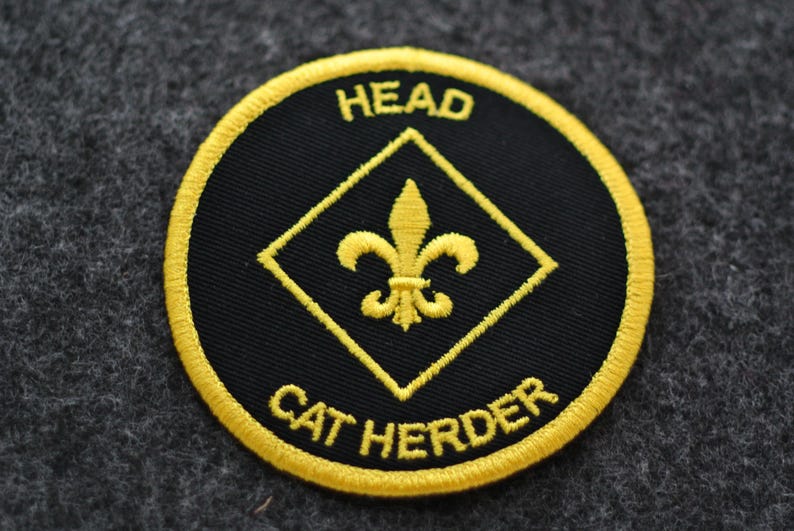 May include: A round, black patch with yellow embroidered text and accents. The text reads "HEAD" at the top and "CAT HERDER" at the bottom. A yellow fleur-de-lis is centered inside a yellow diamond.