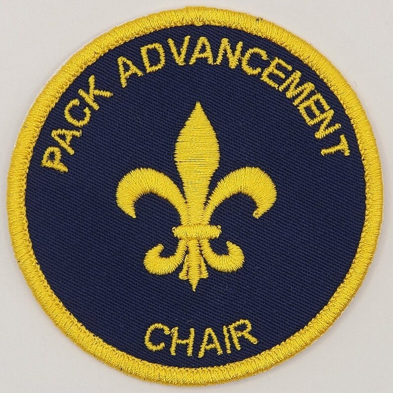 May include: A round, yellow embroidered patch with a blue background. The patch features a fleur-de-lis in the centre and the text "PACK ADVANCEMENT CHAIR" around the edge.