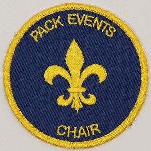 May include: A round, blue and gold embroidered patch with the text "PACK EVENTS CHAIR" and a gold fleur-de-lis in the center.