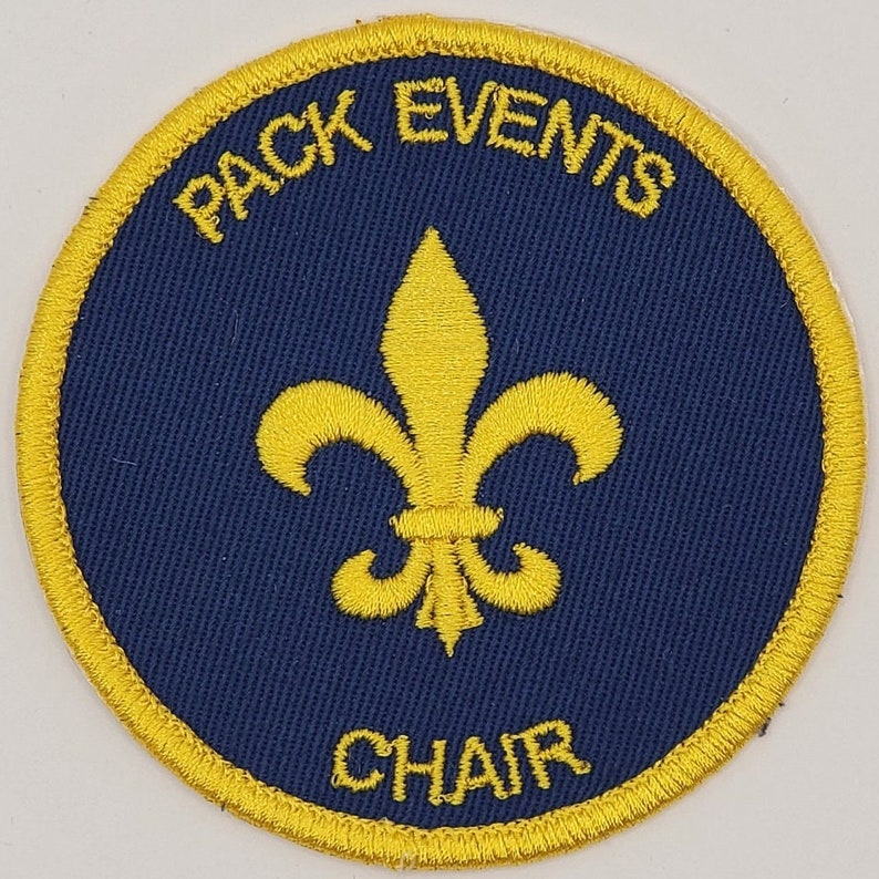 May include: A round, blue and gold embroidered patch with the text "PACK EVENTS CHAIR" and a gold fleur-de-lis in the center.