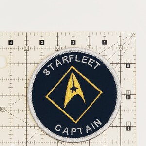 May include: A navy blue round patch with a white border and yellow stitching. The patch features a yellow diamond with a yellow starfleet insignia inside. The text "STARFLEET" is written in white above the diamond and "CAPTAIN" is written in white below the diamond.