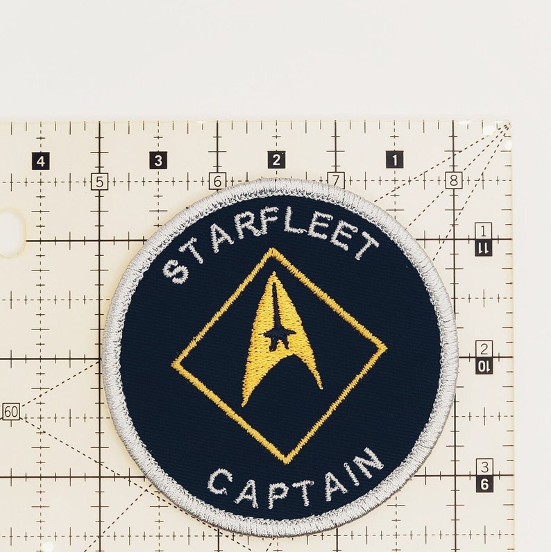 May include: A navy blue round patch with a white border and yellow stitching. The patch features a yellow diamond with a yellow starfleet insignia inside. The text "STARFLEET" is written in white above the diamond and "CAPTAIN" is written in white below the diamond.