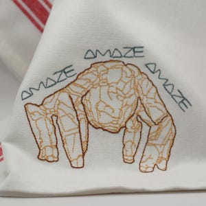 Pu&ograve; includere: Tovagliolo di stoffa bianco con un disegno ricamato di una creatura con un contorno marrone strutturato. La parola "AMAZE" &egrave; ricamata in verde acqua sopra e ai lati della creatura.