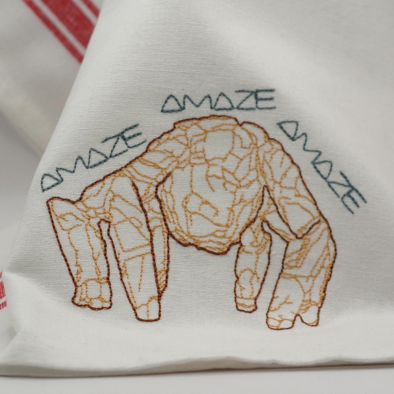 Pu&ograve; includere: Tovagliolo di stoffa bianco con un disegno ricamato di una creatura con un contorno marrone strutturato. La parola "AMAZE" &egrave; ricamata in verde acqua sopra e ai lati della creatura.