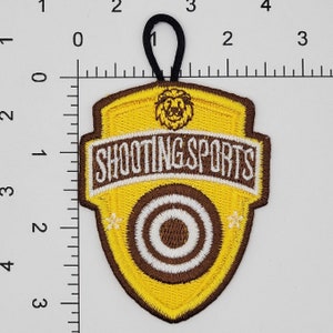 Pu&ograve; includere: Toppa ricamata gialla e marrone con un leone, un bersaglio e il testo "SHOOTING SPORTS". La toppa ha un anello in cima per il fissaggio.