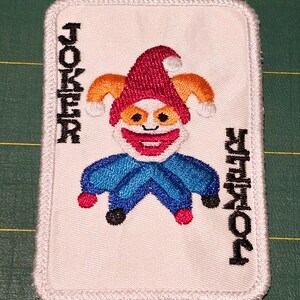Op de afbeelding: Witte geborduurde patch van een joker speelkaart met een rode, oranje en blauwe nar. Het woord "JOKER" is verticaal aan beide kanten van de nar geborduurd.