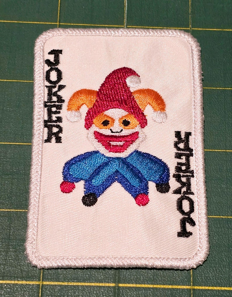 Op de afbeelding: Witte geborduurde patch van een joker speelkaart met een rode, oranje en blauwe nar. Het woord "JOKER" is verticaal aan beide kanten van de nar geborduurd.