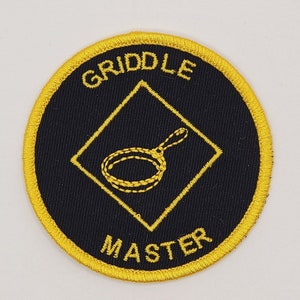 May include: A black and yellow embroidered patch with a diamond shape in the center. The diamond has a yellow outline and a yellow image of a frying pan inside. The text "GRILL MASTER" is written in yellow around the diamond.