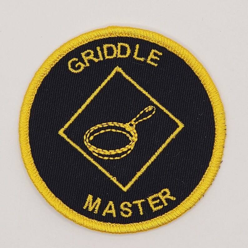 May include: A black and yellow embroidered patch with a diamond shape in the center. The diamond has a yellow outline and a yellow image of a frying pan inside. The text "GRILL MASTER" is written in yellow around the diamond.