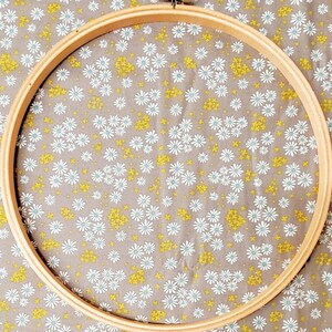 May include: A wooden embroidery hoop with a natural wood finish. The hoop is round and has a diameter of approximately 10 inches. The hoop is lying on a fabric with a white daisy pattern.