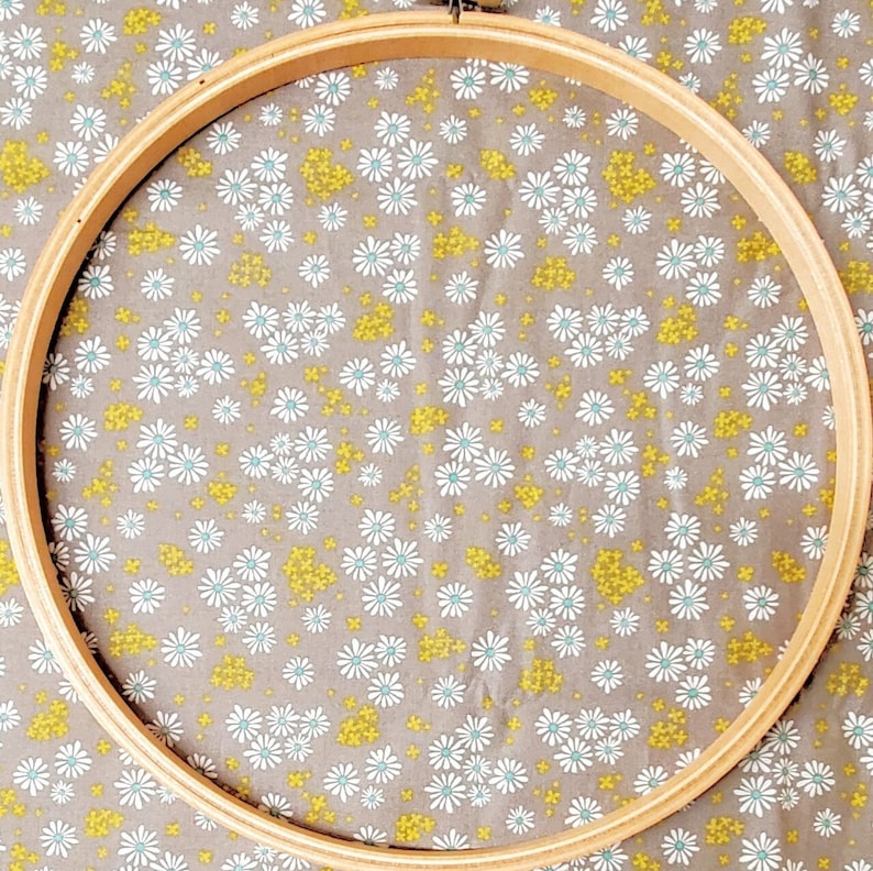 May include: A wooden embroidery hoop with a natural wood finish. The hoop is round and has a diameter of approximately 10 inches. The hoop is lying on a fabric with a white daisy pattern.