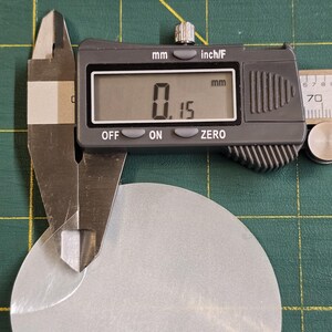 May include: A digital caliper measuring the thickness of a silver-colored, oval-shaped metal piece. The digital display shows a measurement of 0.15 mm. The background is a green cutting mat.