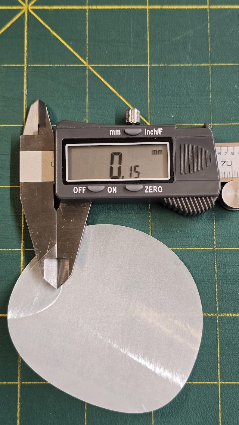 May include: A digital caliper measuring the thickness of a silver-colored, oval-shaped metal piece. The digital display shows a measurement of 0.15 mm. The background is a green cutting mat.