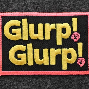 May include: Embroidered patch with the word "GLURP!" in yellow, stacked on a black background, framed by a pink border. Two pink skull graphics are on the right side of the patch.