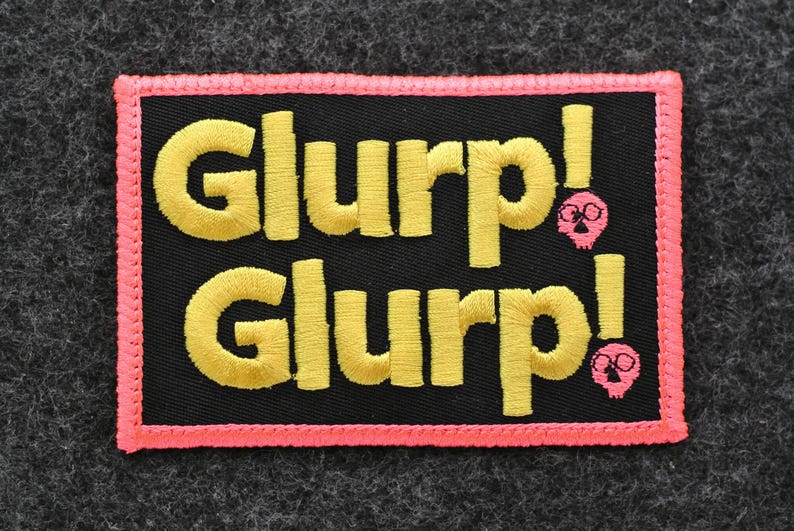 May include: Embroidered patch with the word "GLURP!" in yellow, stacked on a black background, framed by a pink border. Two pink skull graphics are on the right side of the patch.