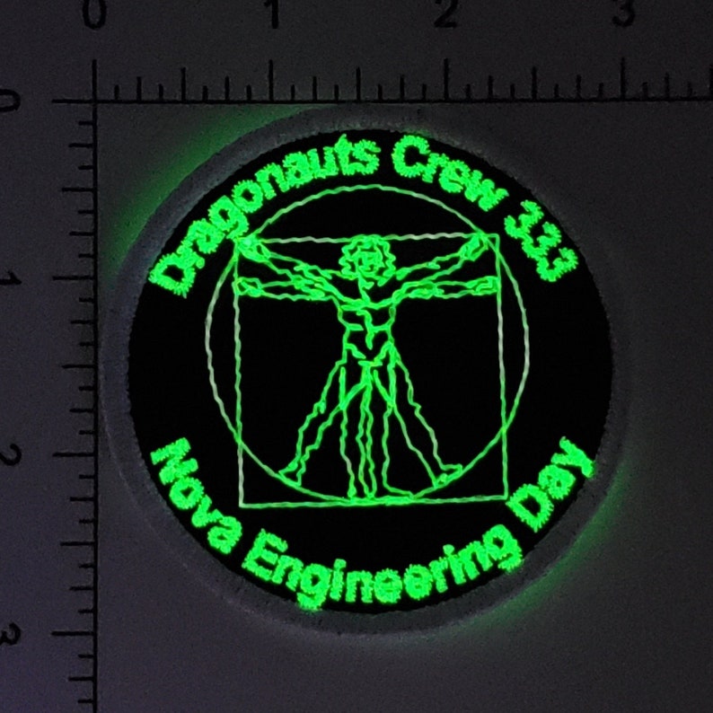 Op de afbeelding: Een zwart en groen geborduurde patch met de tekst "Dragonaunts Crew 33" en "Nova Engineering Day" rond een groene omtrek van een Vitruviusman.
