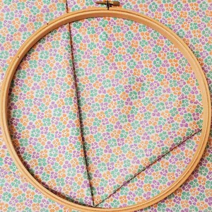 May include: A wooden embroidery hoop holds floral fabric. The fabric features a pattern of small flowers in shades of orange, purple, and green on a white background. The hoop is made of light-coloured wood.