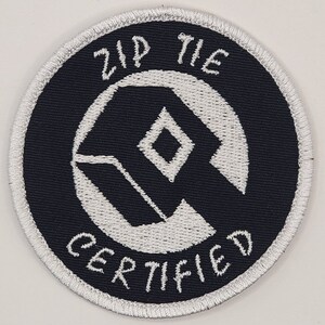 May include: A white embroidered patch with a black background. The patch features a white geometric design with a diamond shape in the center. The text "Zip Tie Certified" is written in a white cursive font around the geometric design.