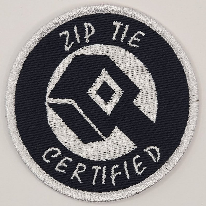 Może przedstawiać: Biała naszywka haftowana na czarnym tle. Naszywka przedstawia biały geometryczny wz&oacute;r z diamentem w środku. Tekst "Zip Tie Certified" jest napisany białym kursywą wok&oacute;ł wzoru geometrycznego.