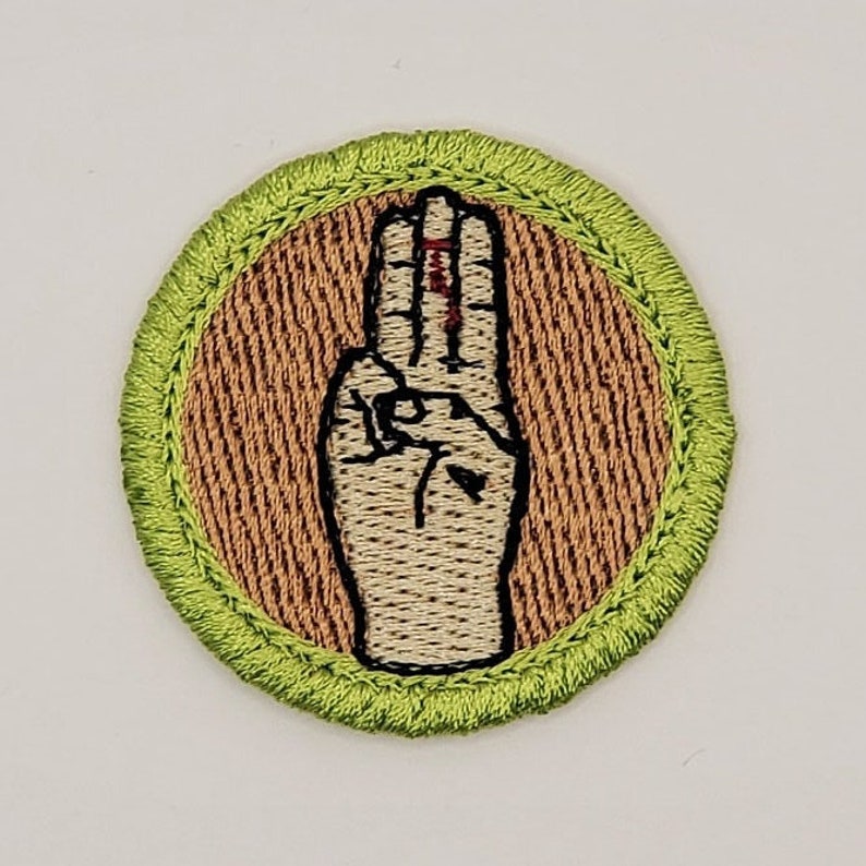 May include: A green and brown embroidered scout badge with a hand making the scout sign.