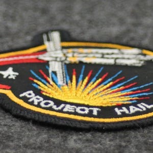 Puede incluir: Parche bordado con fondo negro, con una nave espacial y el texto "PROJECT HAIL MARY". El parche tiene una combinaci&oacute;n de colores amarillo, rojo y azul, con las palabras "PROJECT HAIL MARY" y "MARY" en blanco.