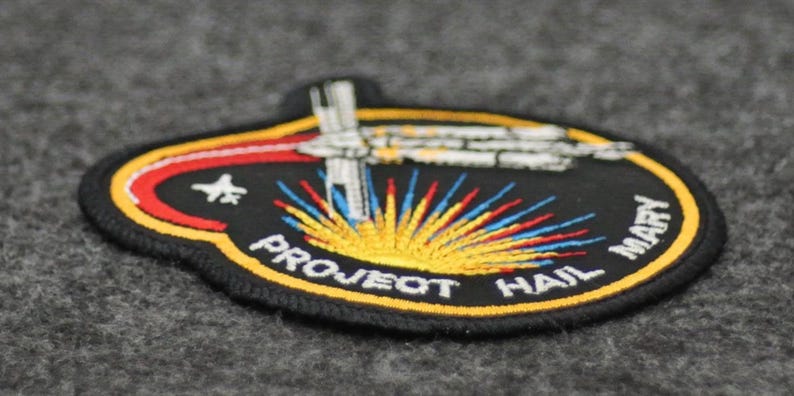 Puede incluir: Parche bordado con fondo negro, con una nave espacial y el texto "PROJECT HAIL MARY". El parche tiene una combinaci&oacute;n de colores amarillo, rojo y azul, con las palabras "PROJECT HAIL MARY" y "MARY" en blanco.