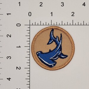 Op de afbeelding: Ronde geborduurde patch met een blauw hamerhaai-ontwerp op een tan achtergrond. De haai is gedetailleerd met witte accenten en de patch heeft een tan rand. De patch heeft een diameter van ongeveer 5 cm.