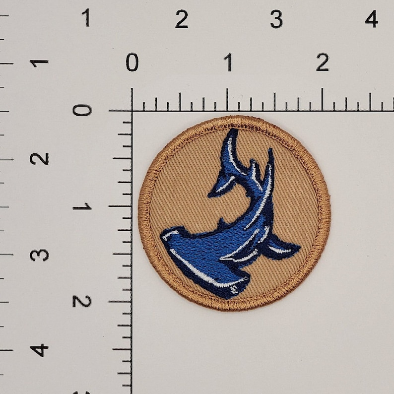 Op de afbeelding: Ronde geborduurde patch met een blauw hamerhaai-ontwerp op een tan achtergrond. De haai is gedetailleerd met witte accenten en de patch heeft een tan rand. De patch heeft een diameter van ongeveer 5 cm.
