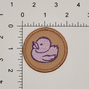 Op de afbeelding: Een bruin geborduurd patch met een paarse rubberen eend op een beige achtergrond.