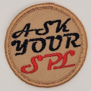 May include: A round brown patch with gold stitching around the edge. The patch has black embroidered text that says "Ask Your" and red embroidered text that says "SPC".