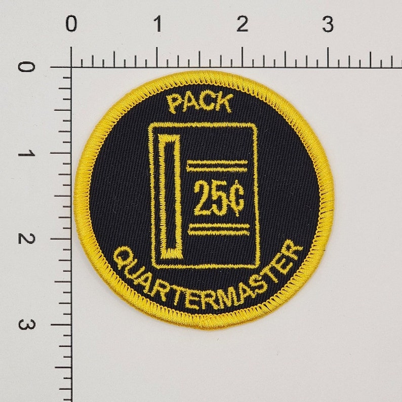 Pode incluir: Um remendo bordado amarelo e preto com o texto "PACK QUARTERMASTER" e uma representa&ccedil;&atilde;o de uma m&aacute;quina de venda autom&aacute;tica com o texto "25&cent;" dentro.