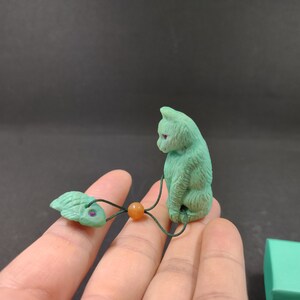 May include: A green jade cat figurine with red eyes, a green jade fish charm, and an orange bead on a black cord.
