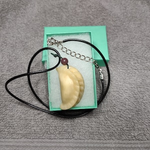 May include: A dumpling-shaped pendant necklace with a black cord, a silver chain, and a small purple bead. The pendant is a light beige color and is presented in a small, open, turquoise gift box.