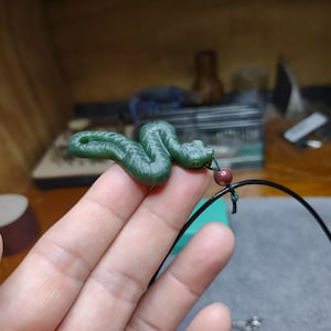 May include: A green snake pendant on a black cord with a red bead. The pendant is made of a smooth, green material and has a detailed snake design.
