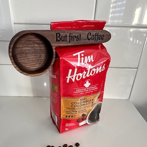 Op de afbeelding: Een houten koffieschep met de tekst "But first... Coffee" bevestigd aan een zak Tim Hortons 100% Colombiaanse koffie. De koffiezak is rood met een wit esdoornbladlogo. Op de voorgrond ligt een hoop koffiebonen.