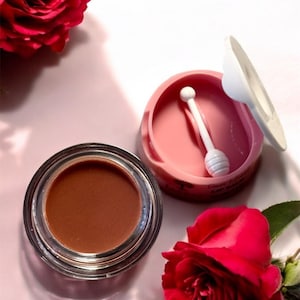 May include: A small round jar of brown lip gloss with a white applicator. The jar is open and the applicator is inside. There are two red roses on either side of the jar.