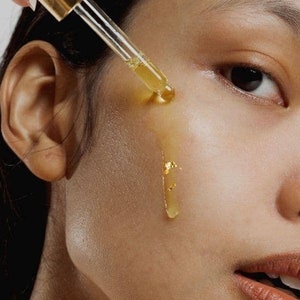 Puede incluir: Primer plano de la cara de una persona con un gotero de aceite facial dorado que se aplica en la mejilla. El aceite gotea por la mejilla y hay peque&ntilde;os trozos de pan de oro en la piel.
