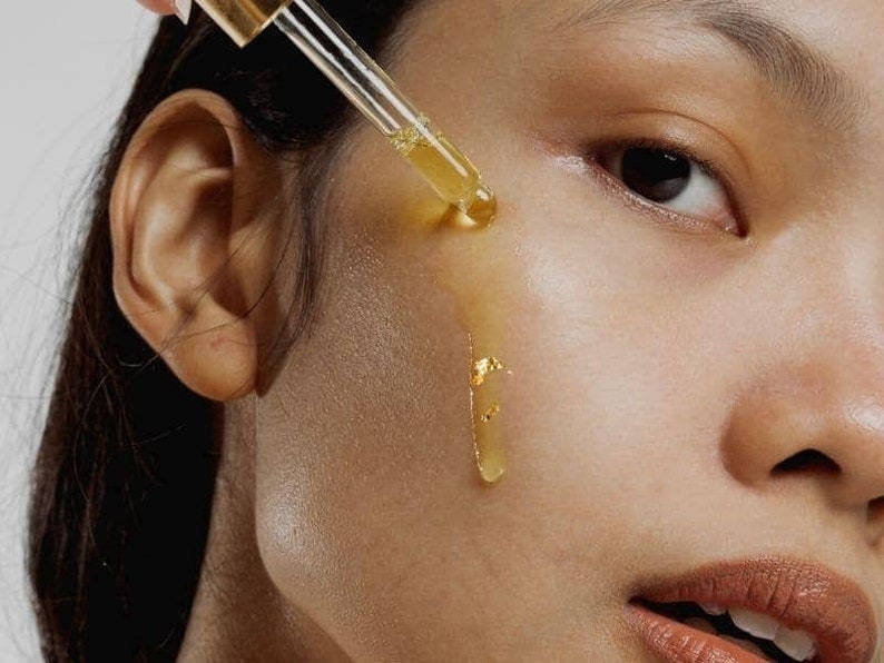 Puede incluir: Primer plano de la cara de una persona con un gotero de aceite facial dorado que se aplica en la mejilla. El aceite gotea por la mejilla y hay peque&ntilde;os trozos de pan de oro en la piel.